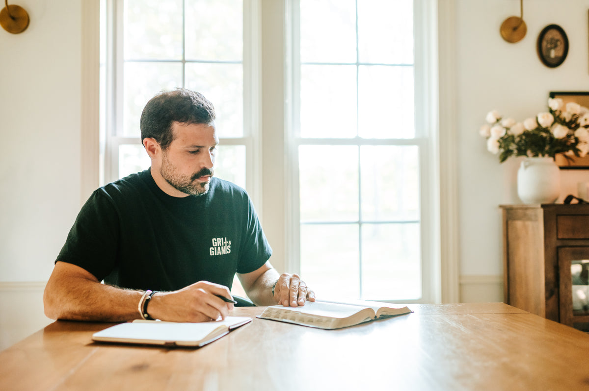 Man wearing a black Grit & Giants shirt while reading the Bible and writing at a table.