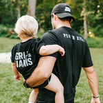 Father carrying his young son while they wear black Grit & Giants shirts outdoors.