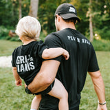 Father carrying his young son while they wear black Grit & Giants shirts outdoors.