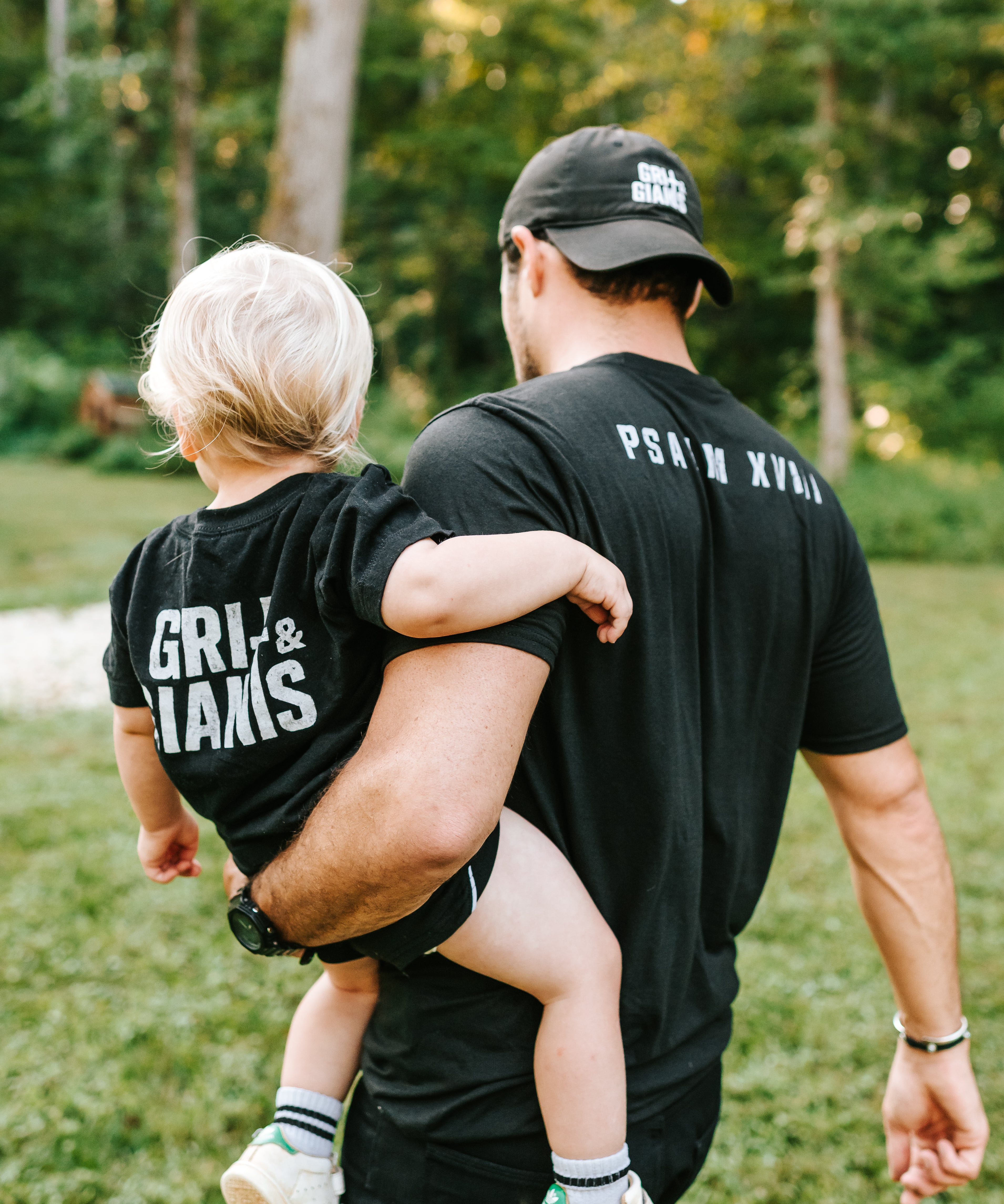 Father carrying his young son while they wear black Grit & Giants shirts outdoors.