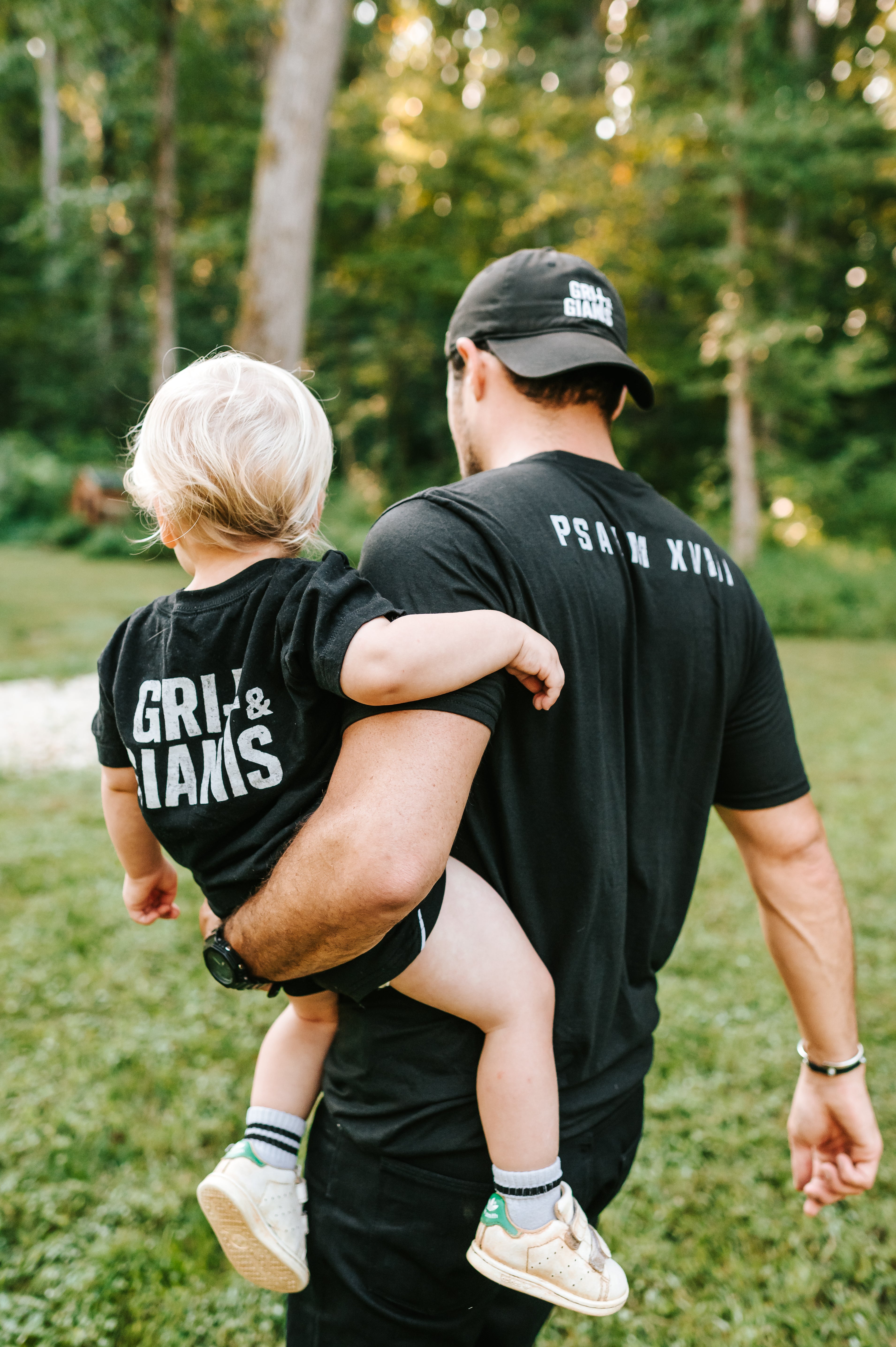 Father carrying his young son while they wear black Grit & Giants shirts outdoors.