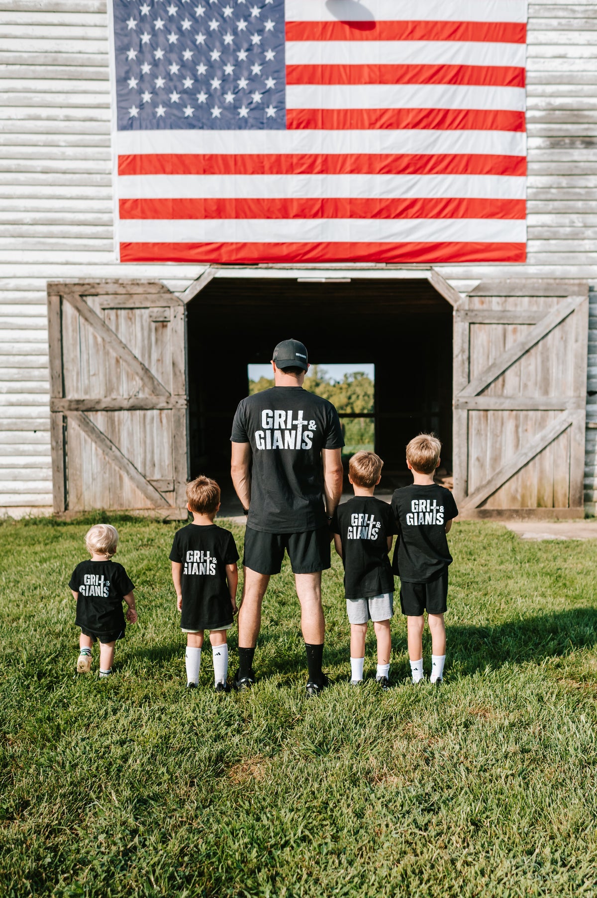 Father and four sons wearing black Grit & Giants shirts in front of a barn and American flag.