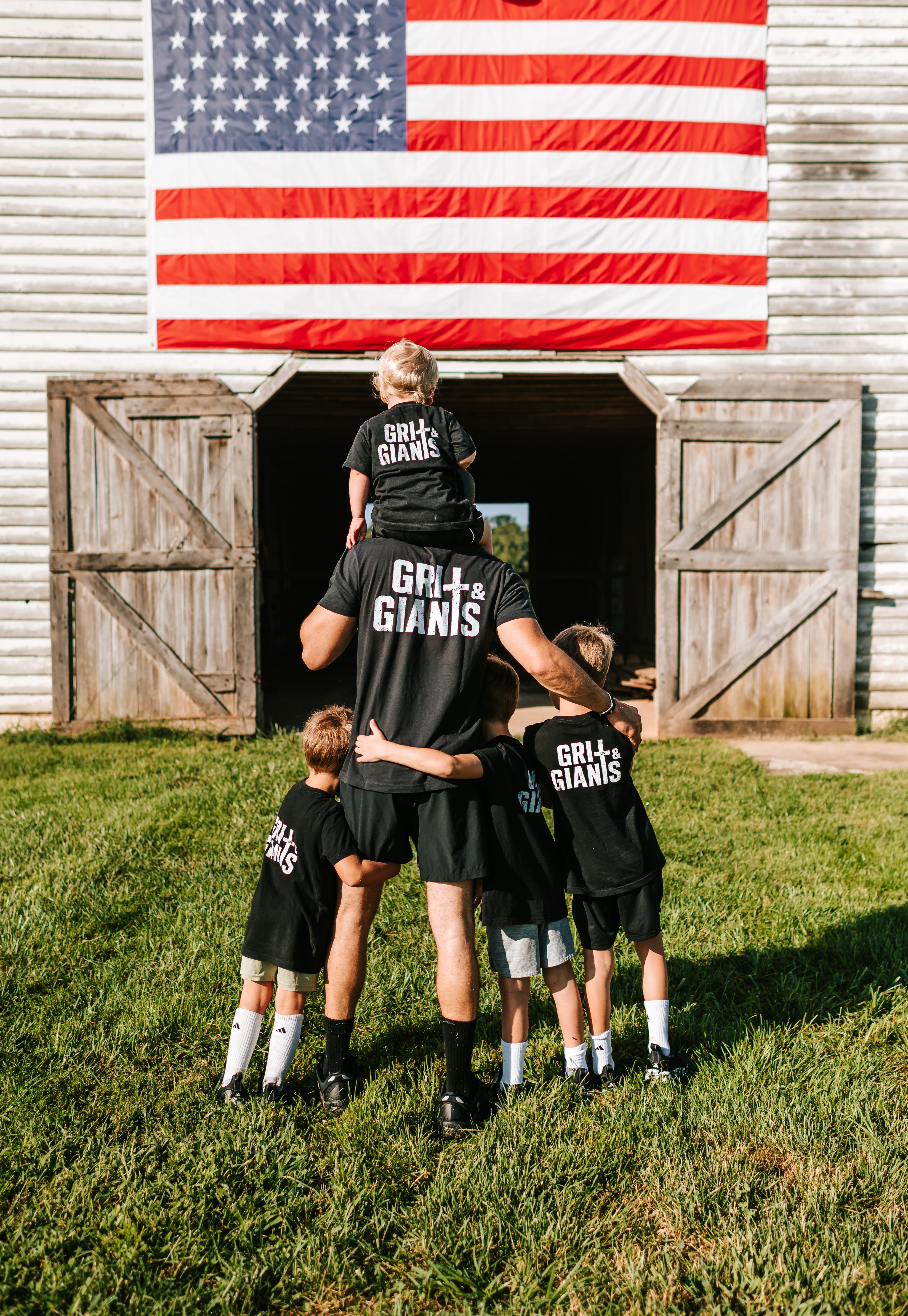 Father and four sons wearing black Grit & Giants shirts in front of a barn and American flag.