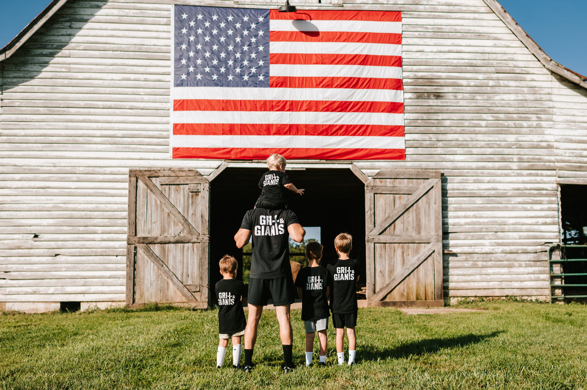 Father and sons wearing Grit & Giants apparel in front of an American flag and barn.