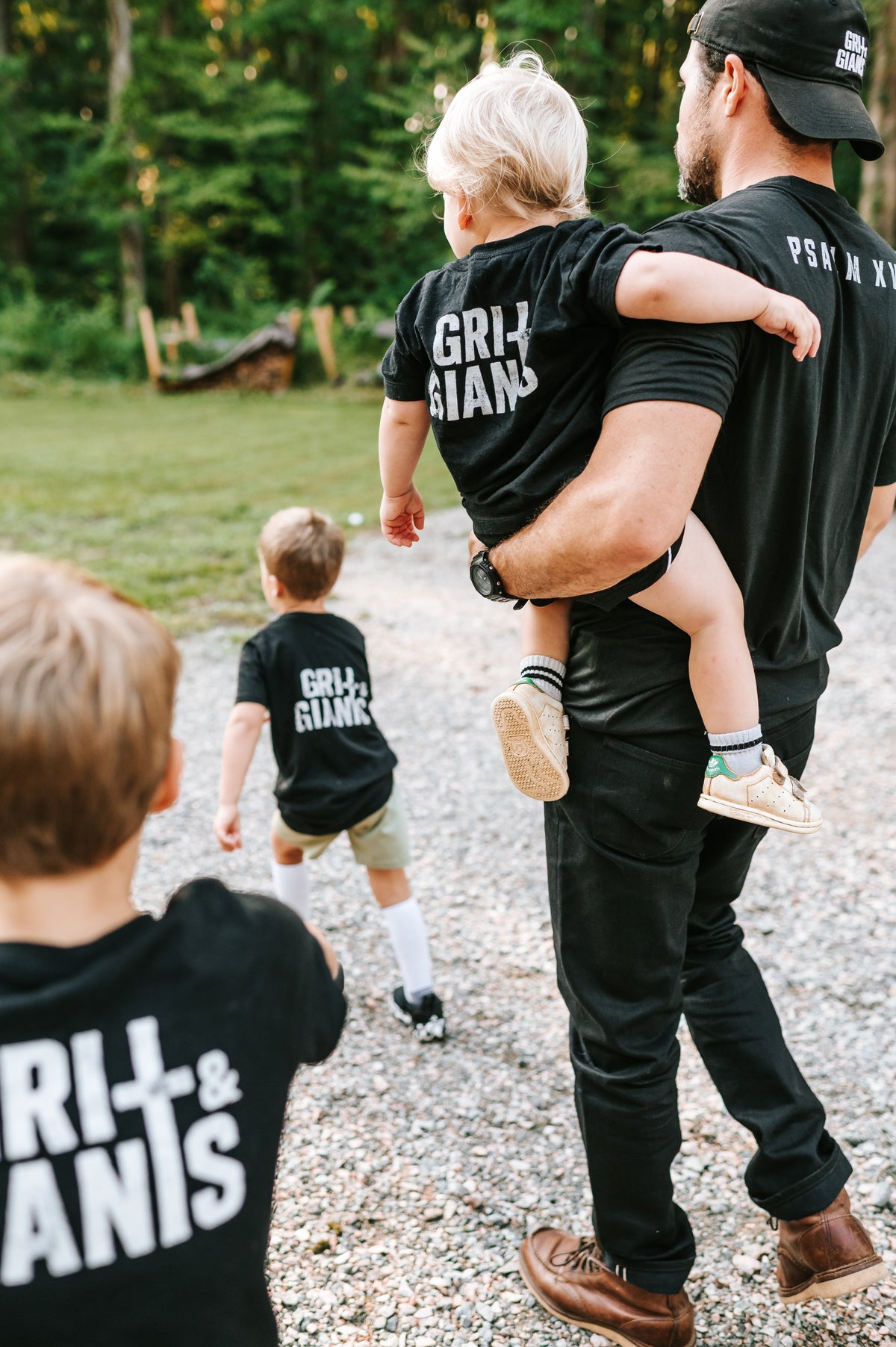 Father carrying his young son while walking outdoors with his boys in matching black Grit & Giants shirts.
