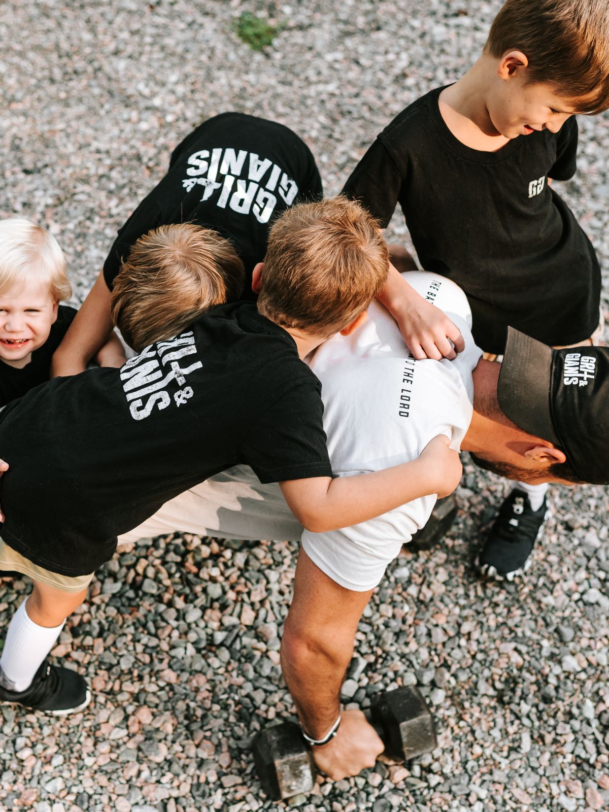 Four boys in black Grit & Giants shirts watching their father lift dumbbells outdoors in a white Grit & Giants tee.