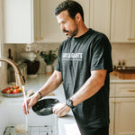 Man wearing a Faded Black Grit & Giants Heavyweight Tee while washing a cast iron skillet in the kitchen.