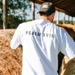Back view of a man wearing a white Grit & Giants Heavyweight Tee featuring Psalm XVIII beside hay bales.