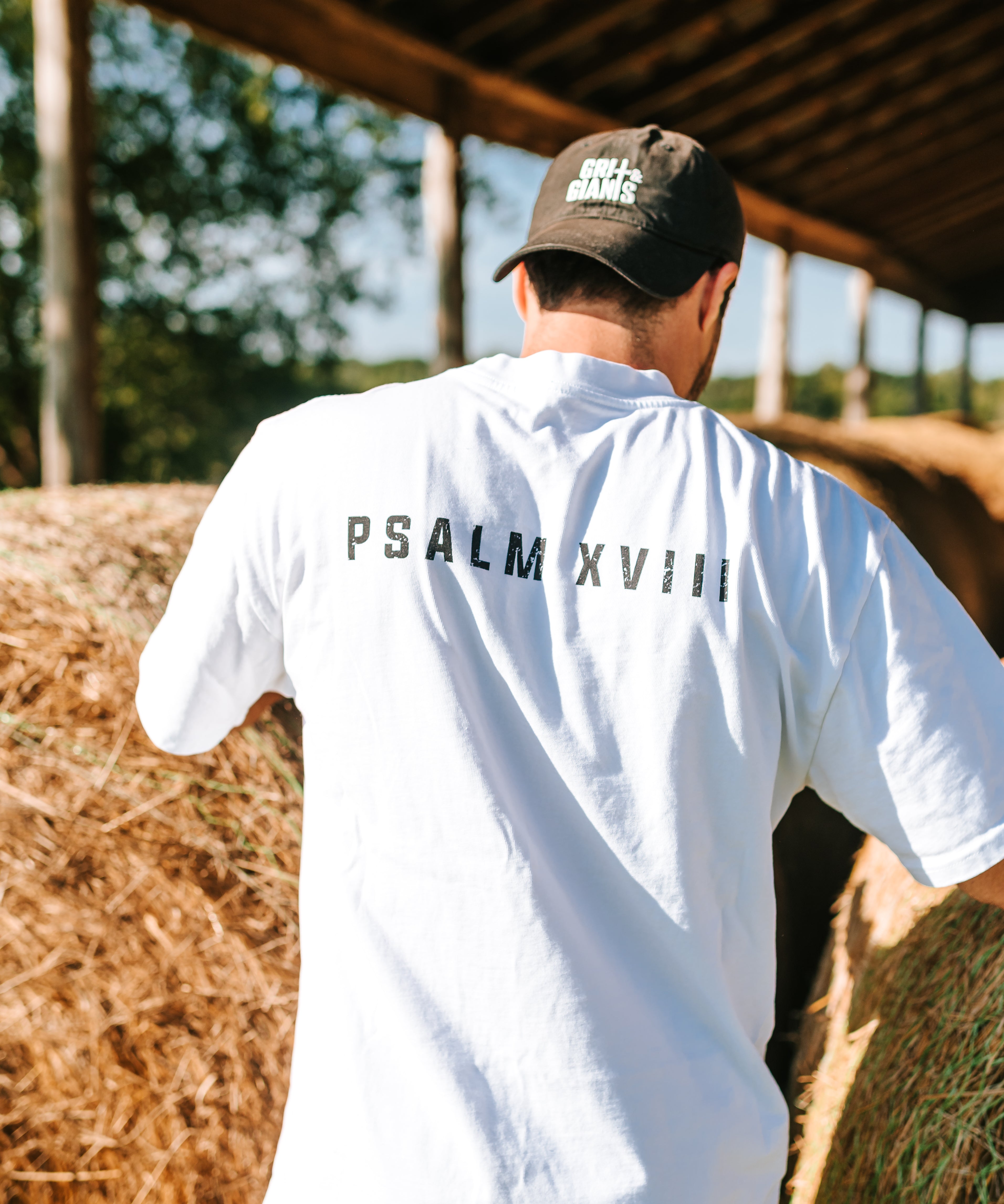 Back view of a man wearing a white Grit & Giants Heavyweight Tee featuring Psalm XVIII beside hay bales.