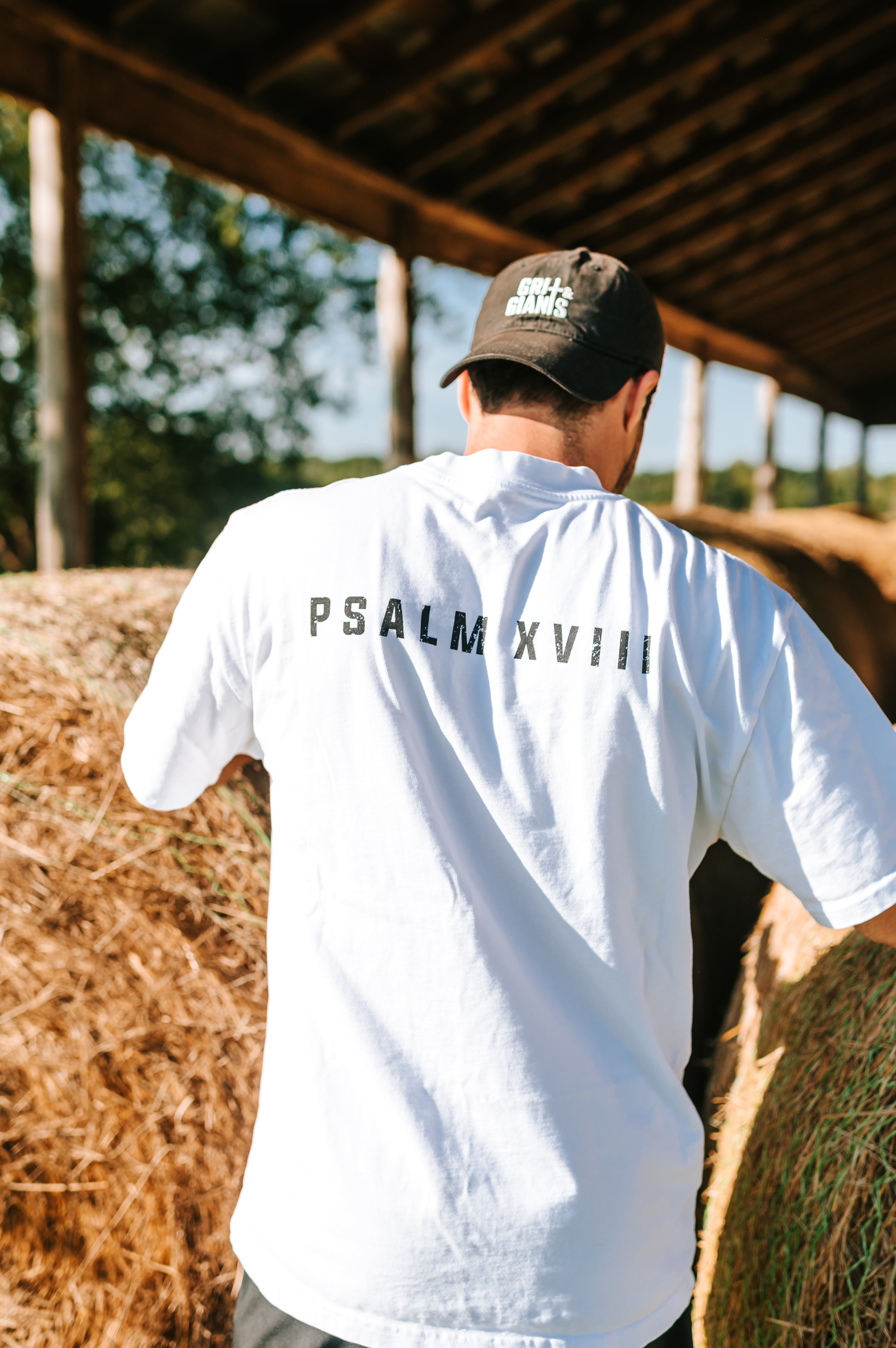 Back view of a man wearing a white Grit & Giants Heavyweight Tee featuring Psalm XVIII beside hay bales.