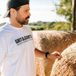 Man wearing a white Grit & Giants Heavyweight Tee outdoors beside hay bales.