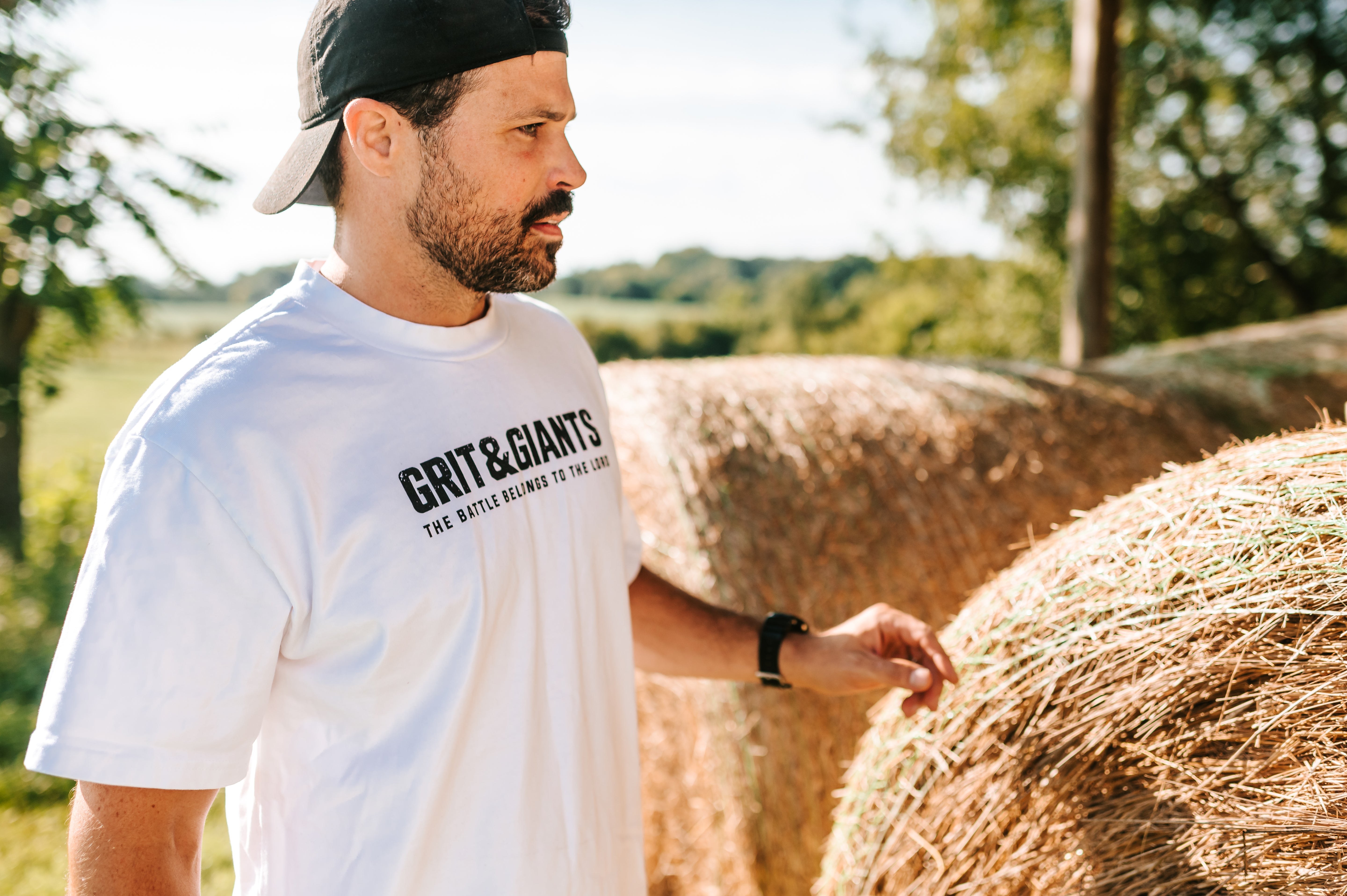 Man wearing a white Grit & Giants Heavyweight Tee outdoors beside hay bales.
