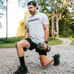 Man wearing the white smoke Grit & Giants Overcomer Training Tee while doing dumbbell lunges outdoors.