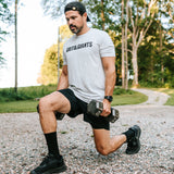 Man wearing the white smoke Grit & Giants Overcomer Training Tee while doing dumbbell lunges outdoors.