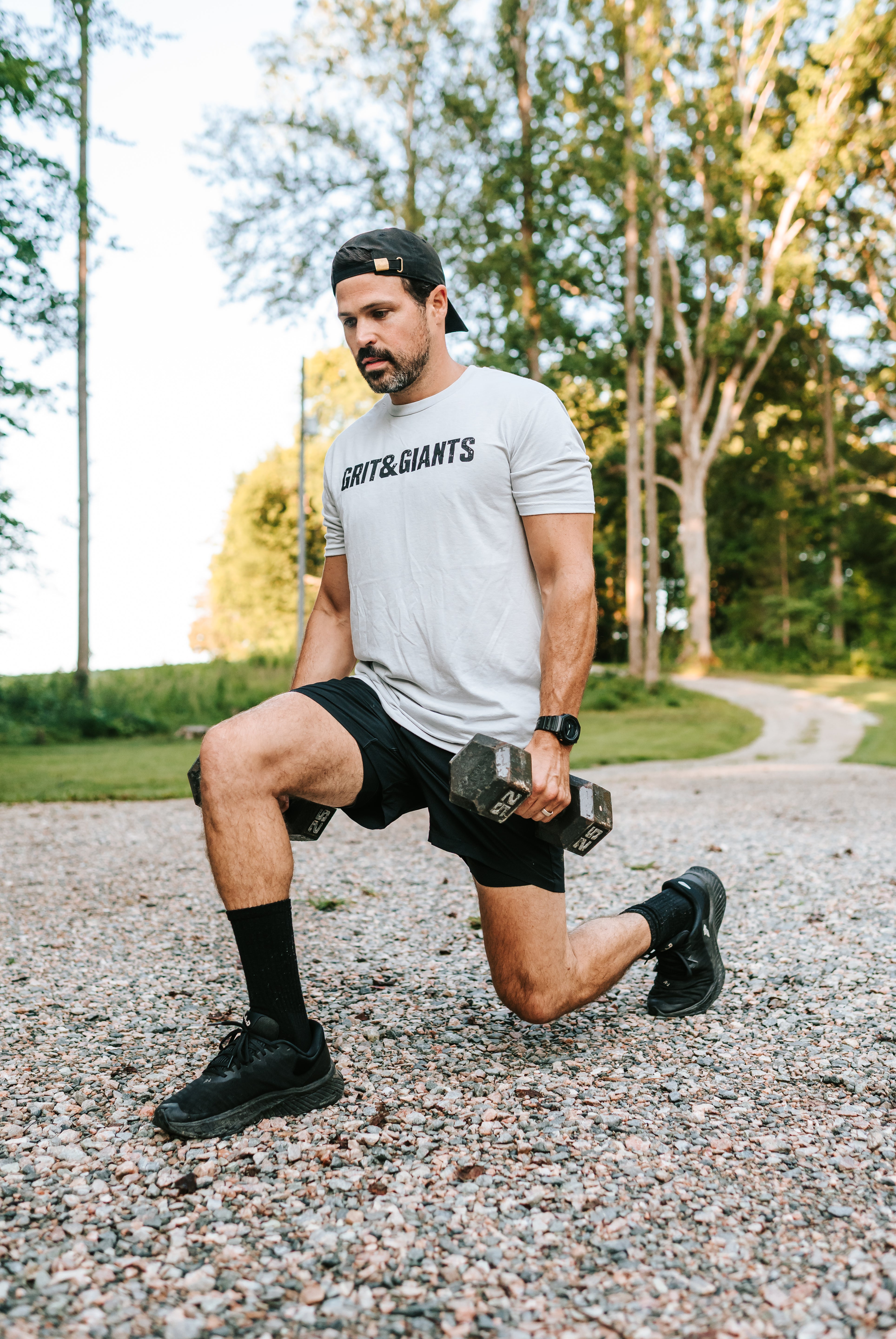 Man wearing the white smoke Grit & Giants Overcomer Training Tee while doing dumbbell lunges outdoors.
