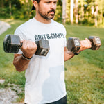 Man wearing the white smoke Grit & Giants Overcomer Training Tee while lifting dumbbells outdoors
