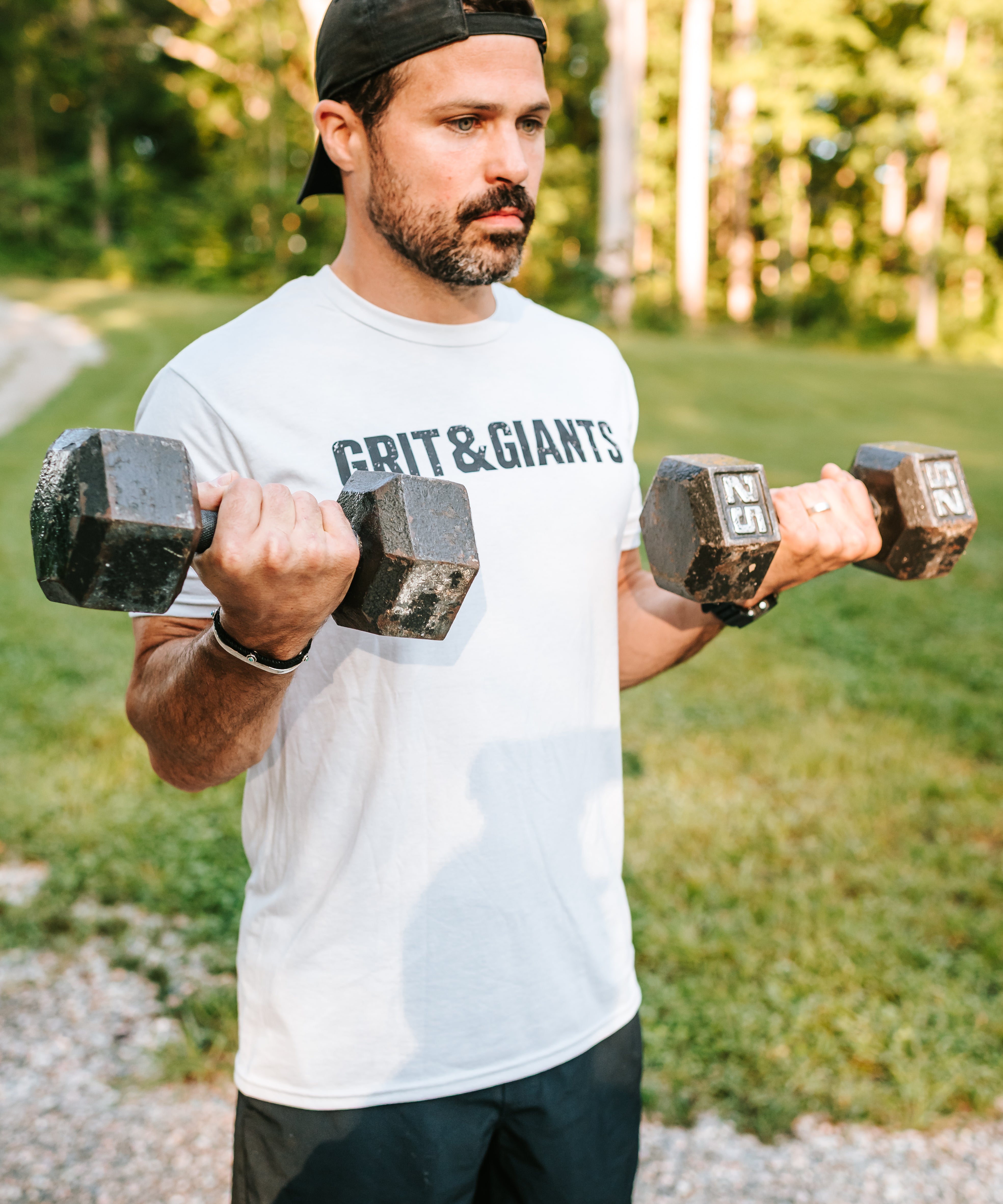 Man wearing the white smoke Grit & Giants Overcomer Training Tee while lifting dumbbells outdoors