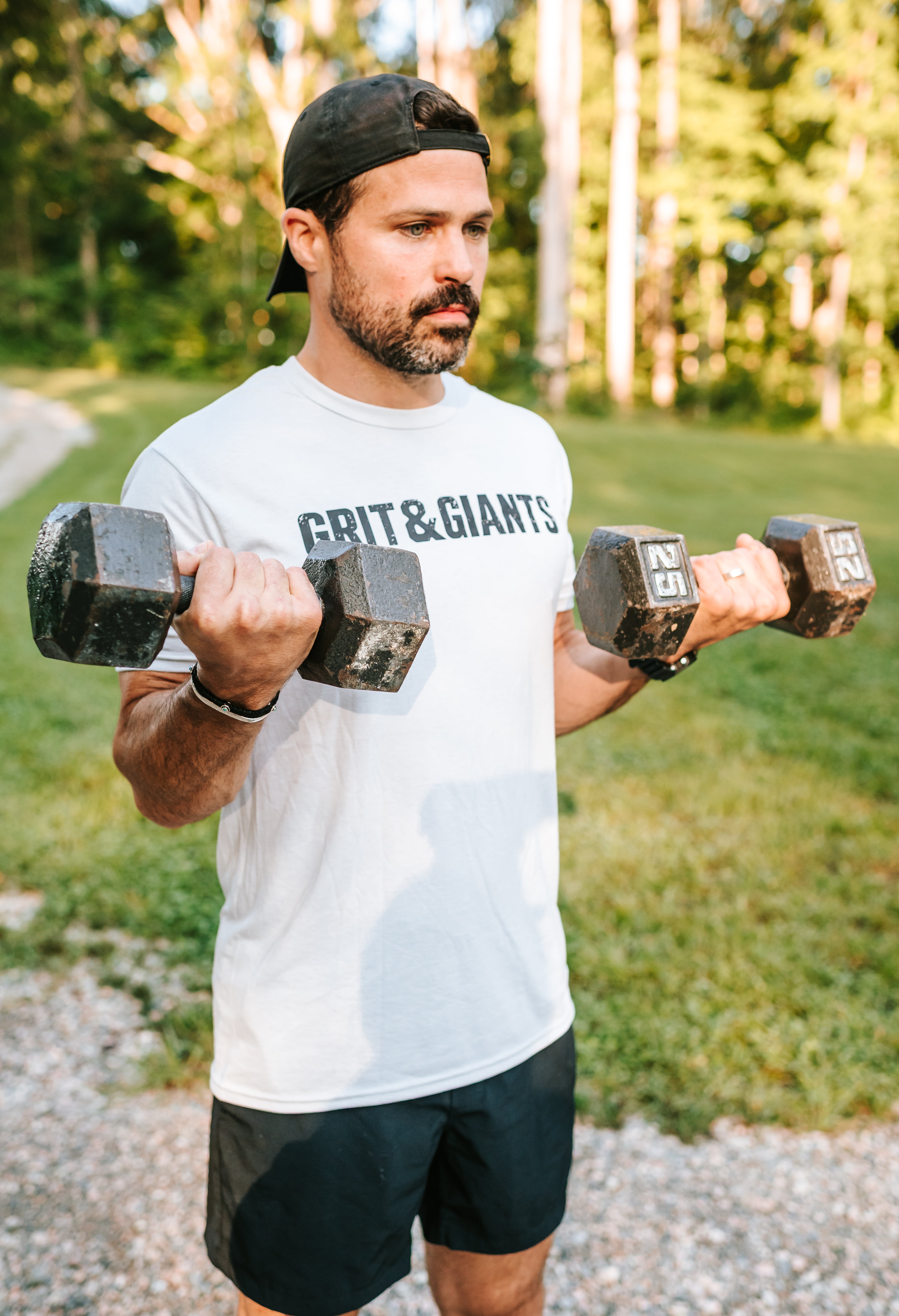 Man wearing the white smoke Grit & Giants Overcomer Training Tee while lifting dumbbells outdoors