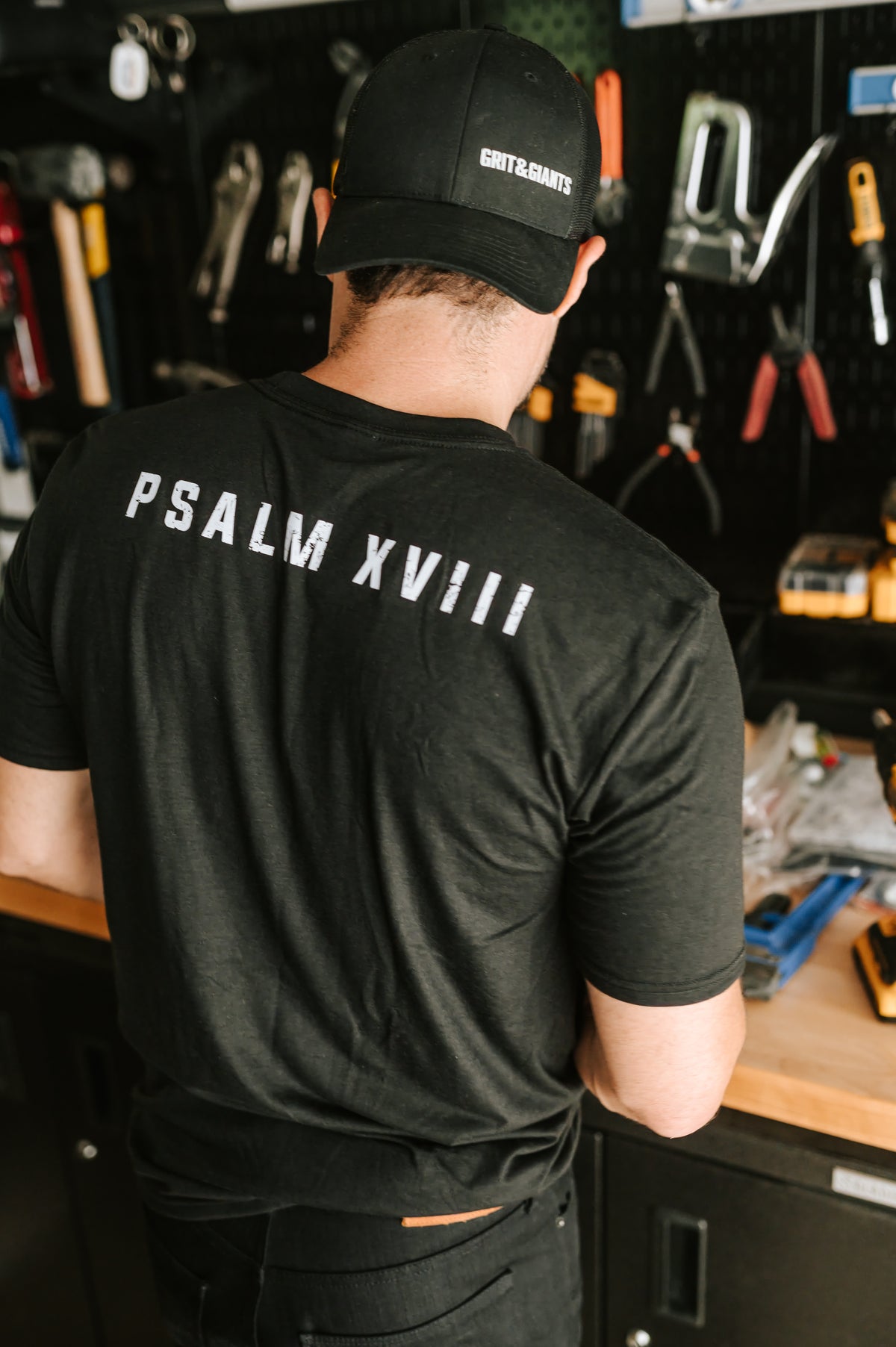 Man wearing a black Grit & Giants Psalm XVIII shirt and cap while working in a garage.