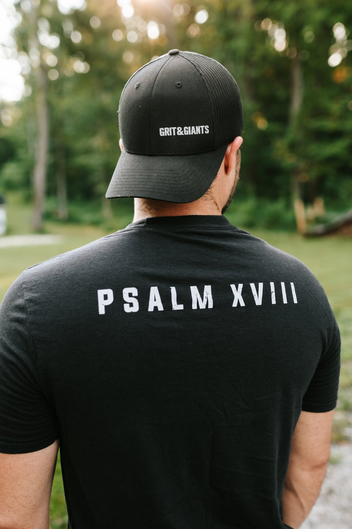 Back view of a man wearing a black Grit & Giants Psalm XVIII training shirt and black cap.