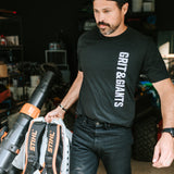 Man wearing a black Grit & Giants Refuge Training Tee while moving equipment in a garage.