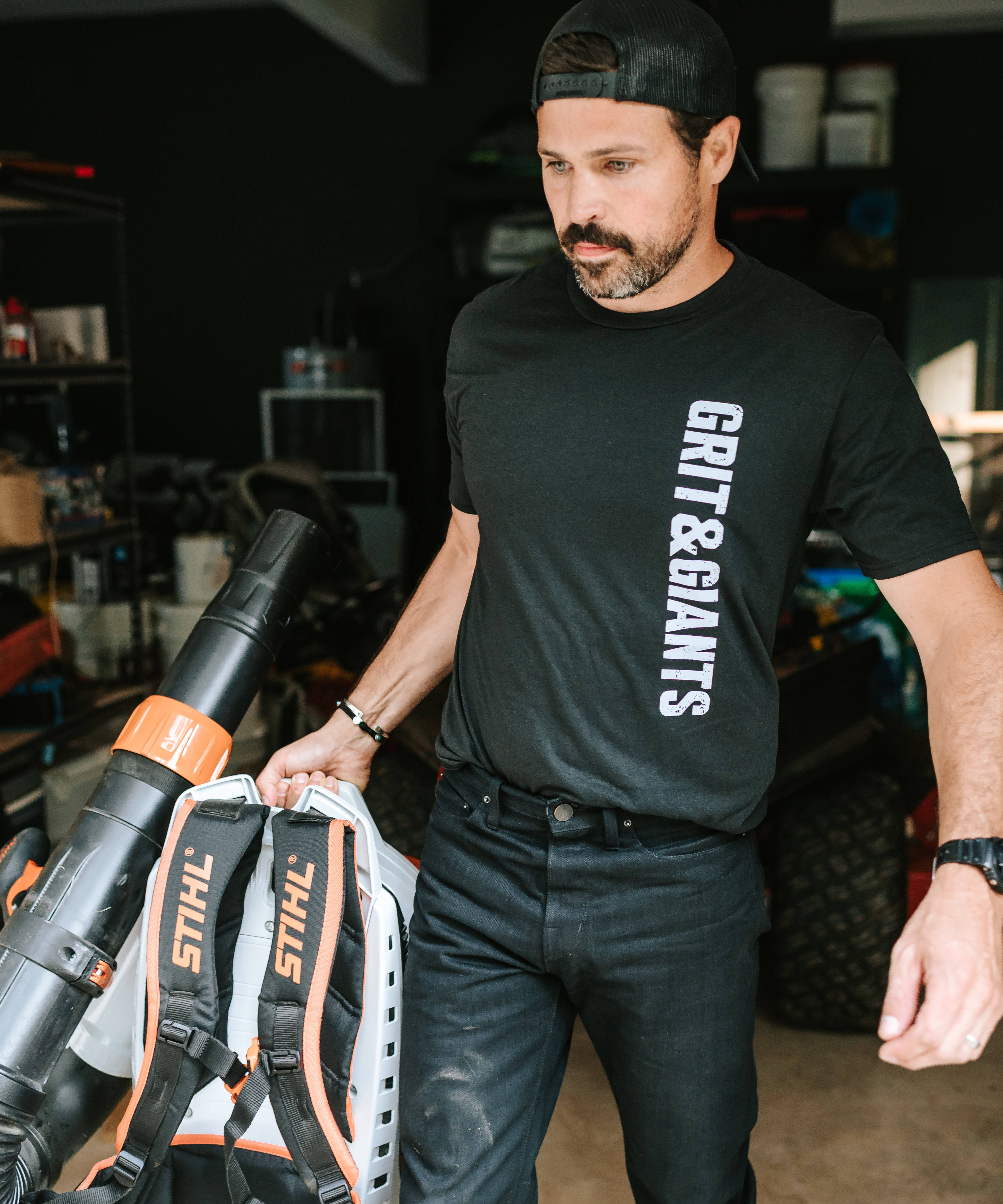 Man wearing a black Grit & Giants Refuge Training Tee while moving equipment in a garage.