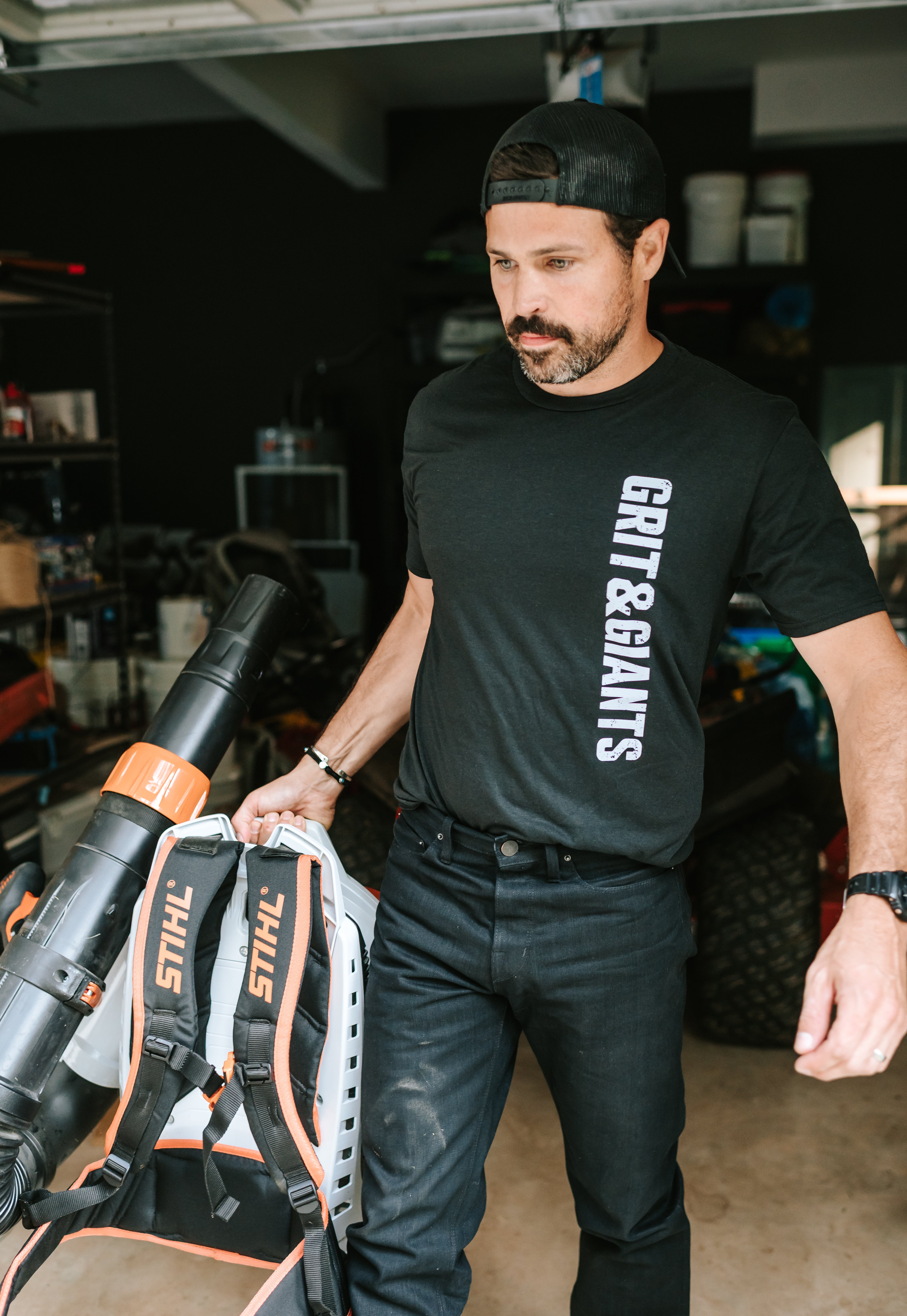 Man wearing a black Grit & Giants Refuge Training Tee while moving equipment in a garage.