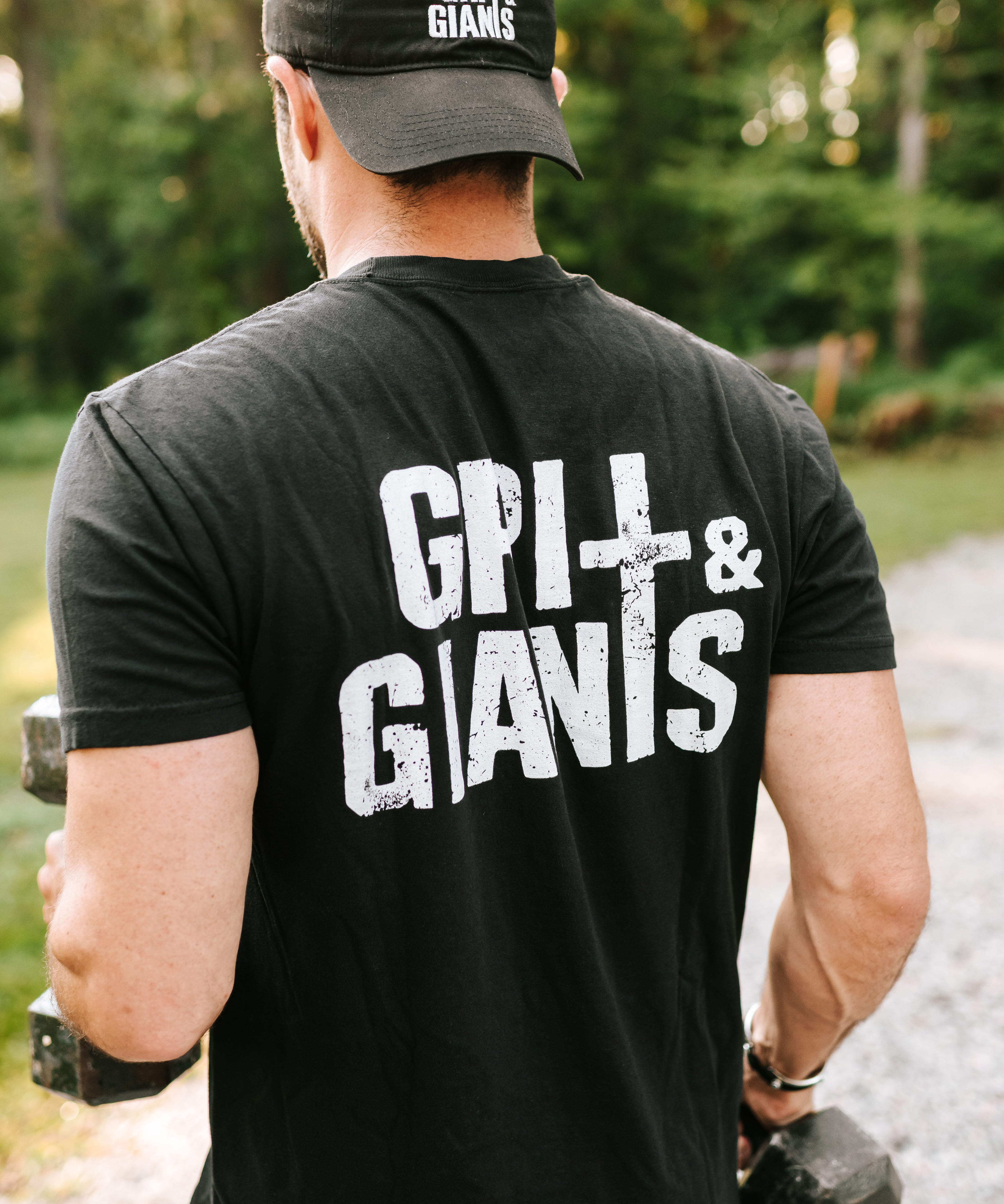 Man lifting dumbbells outdoors wearing a black Grit & Giants Stone Signature Tee with back logo.