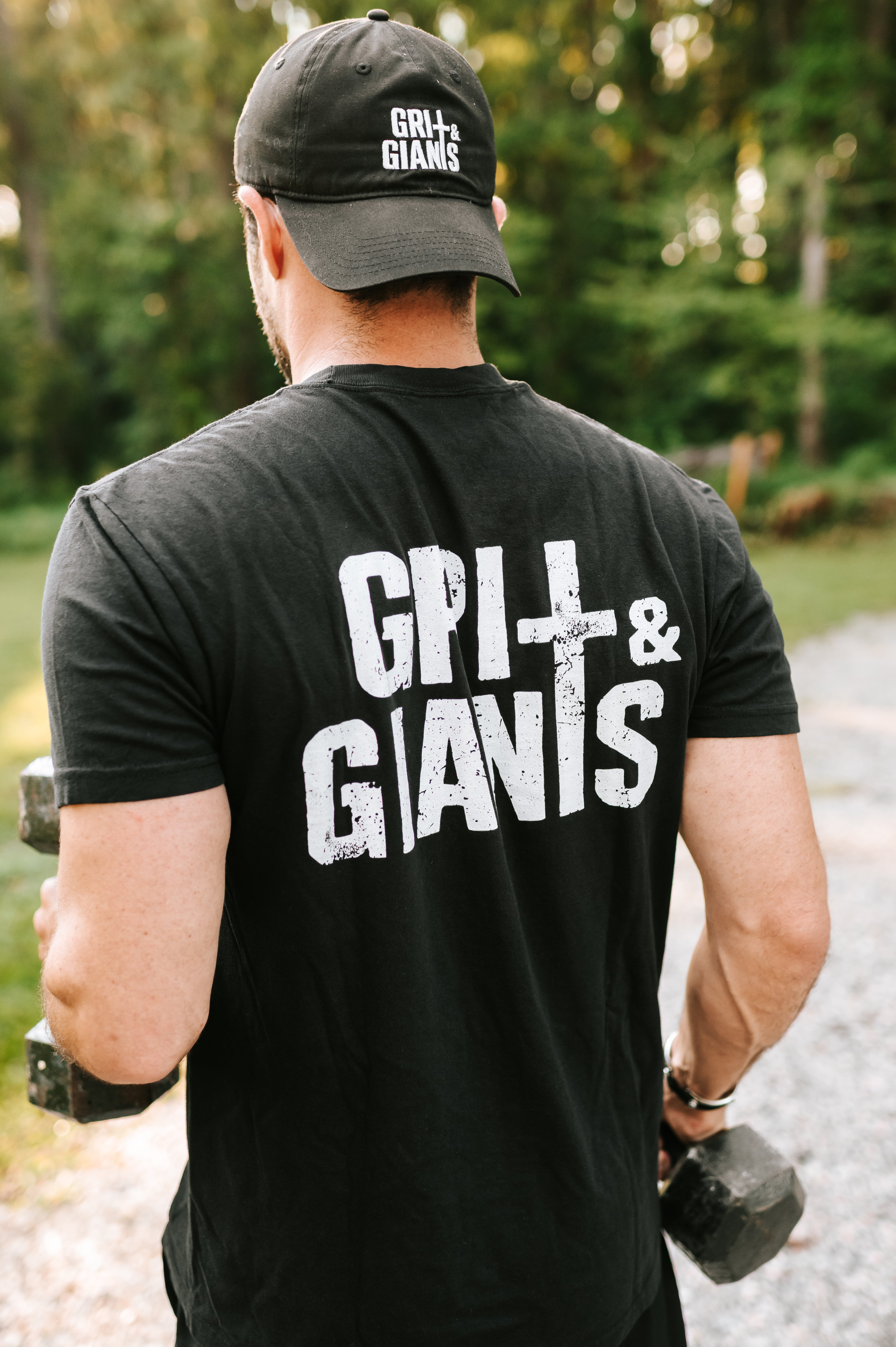 Man lifting dumbbells outdoors wearing a black Grit & Giants Stone Signature Tee with back logo.