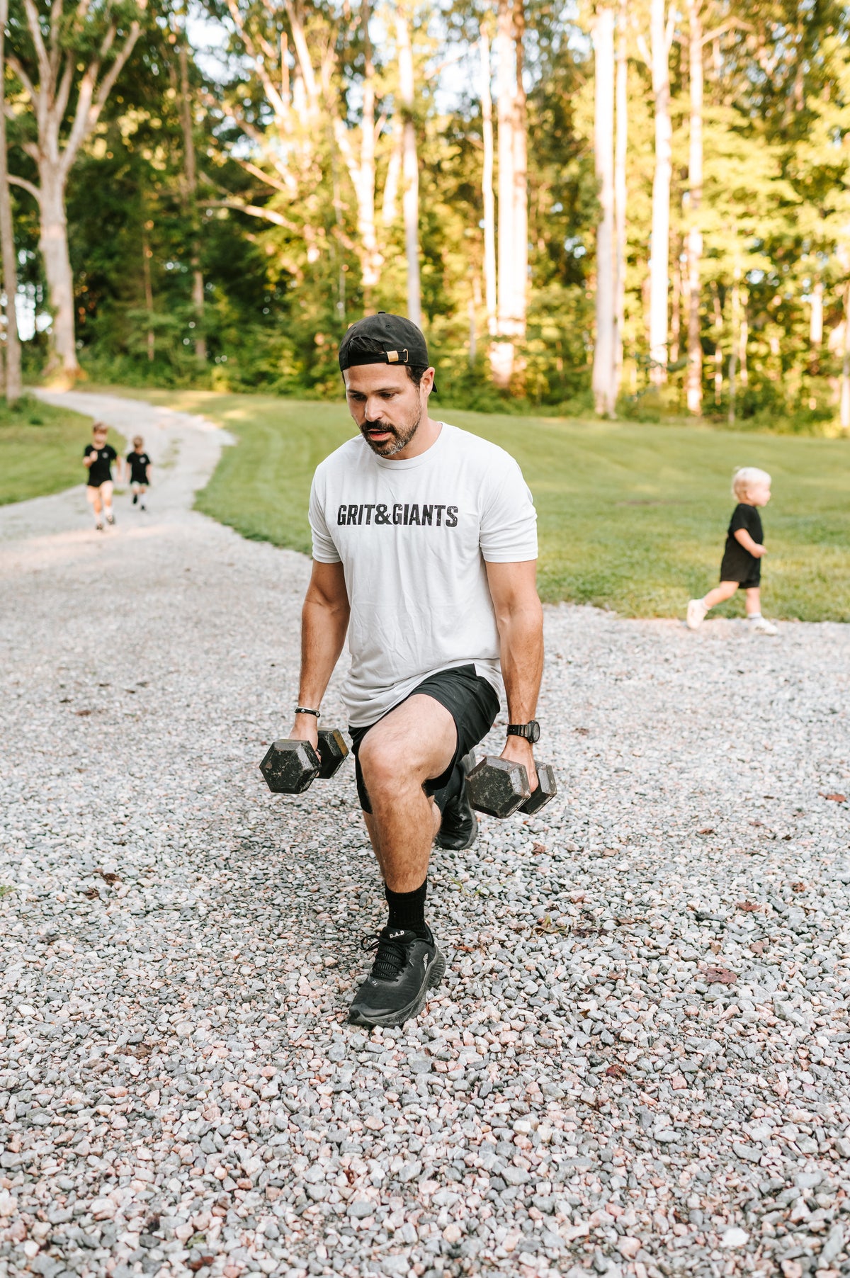 Man wearing a white smoke Grit & Giants training tee doing dumbbell lunges outdoors with his sons in the background.