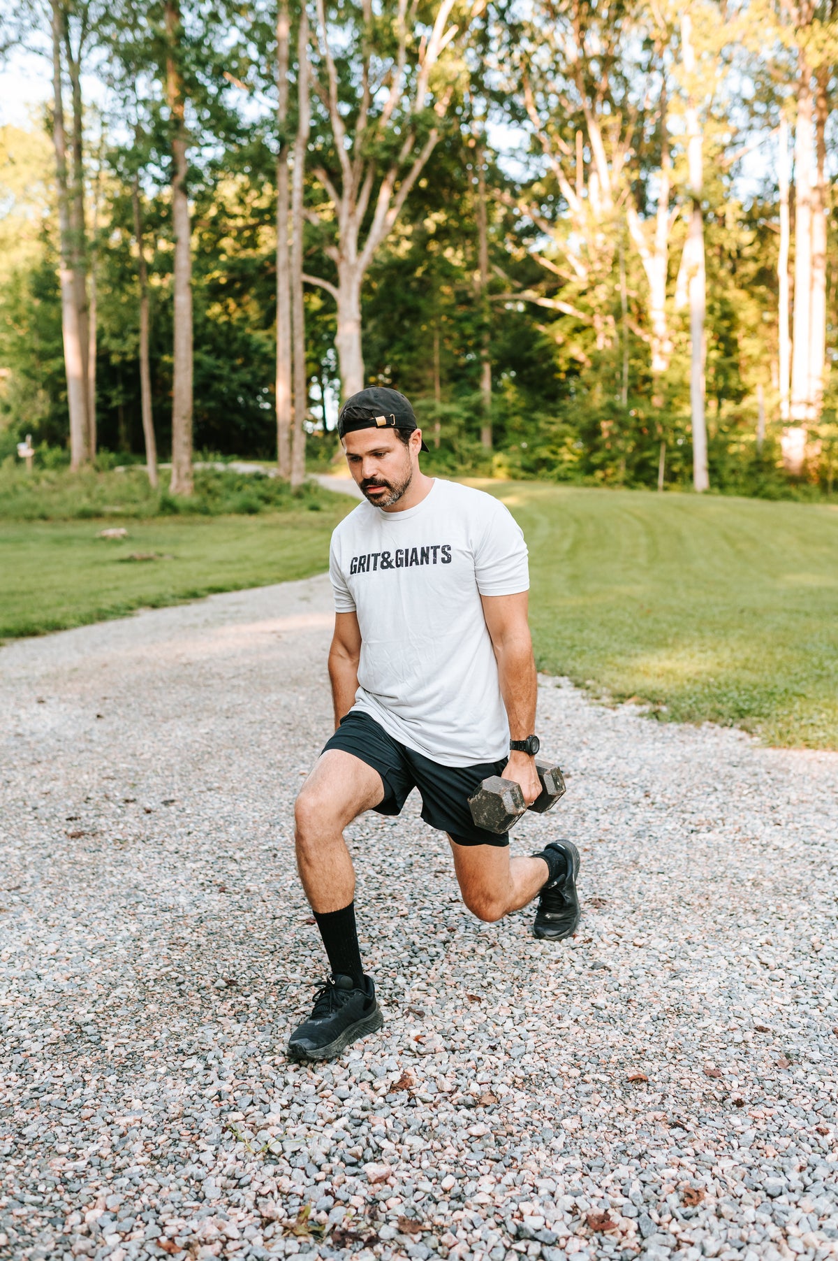 Man wearing a white Grit & Giants training tee while doing dumbbell lunges outdoors.