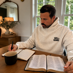 Man wearing a white heather Grit & Giants Victory Hoodie while reading the Bible and writing at a table.