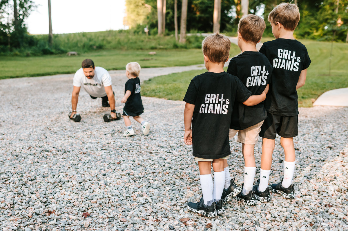 Four boys wearing black Grit & Giants youth shirts while their father works out with dumbbells outdoors.